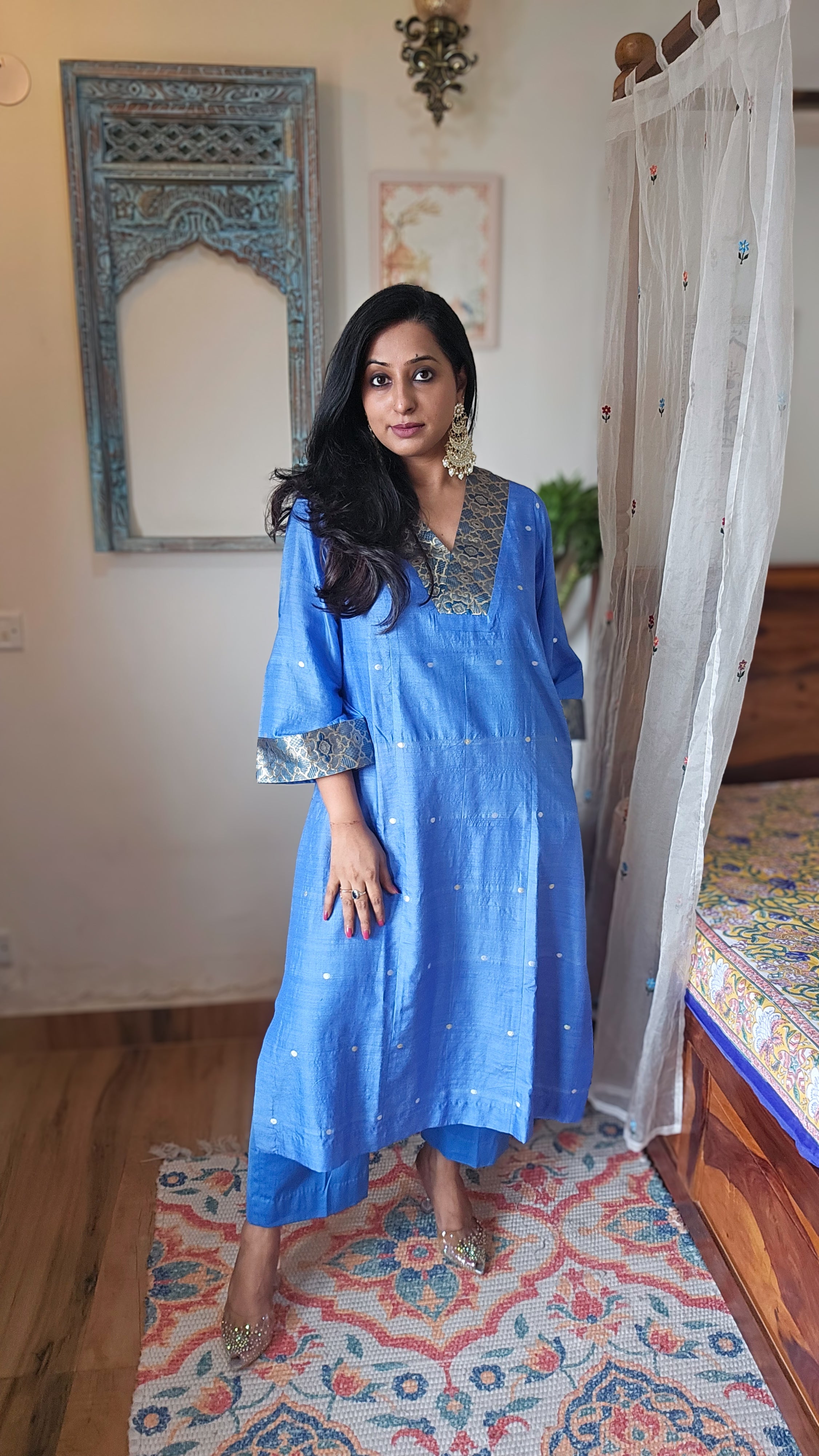 Woman in a bluebanarsi silk traditional outfit standing in a room with a decorative mirror and rug.