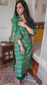 Woman in a green patterned dress standing in a room with wooden furniture and colorful rug.