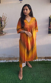 Woman wearing a tie-dye dress standing outdoors with a white wall and greenery in the background