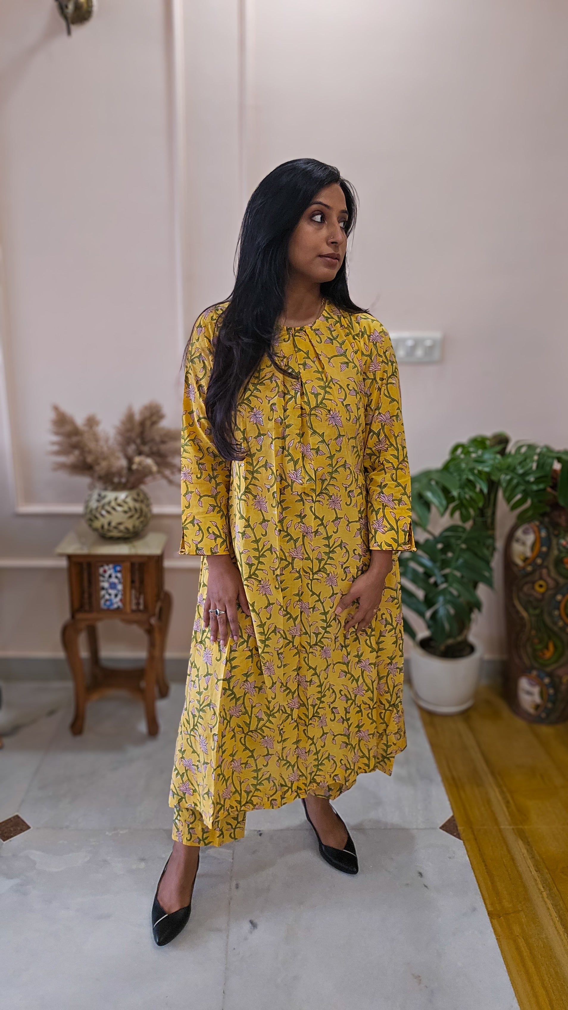 Woman in a yellow floral dress standing in a room with plants and a table.