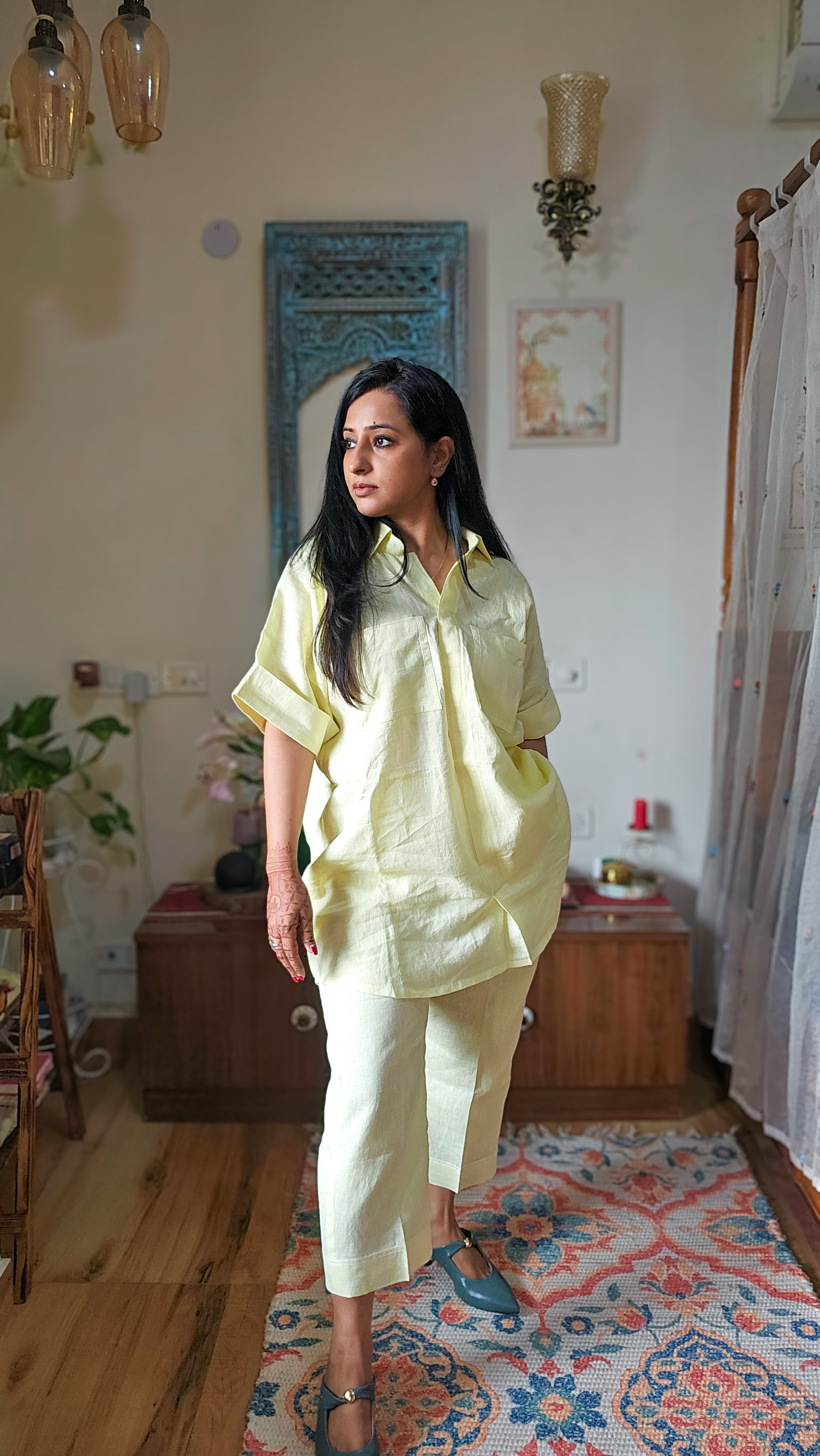 A woman modeling a light yellow linen shirt and matching cropped trousers in a home setting with wooden floors, a patterned rug, and traditional decor.