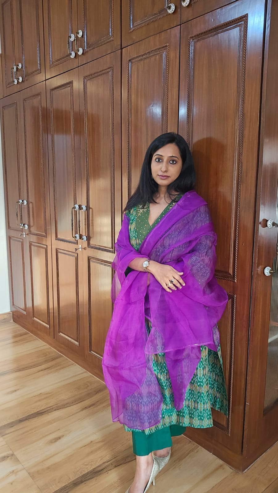 Woman in a purple and green traditional outfit standing in front of wooden wardrobes.