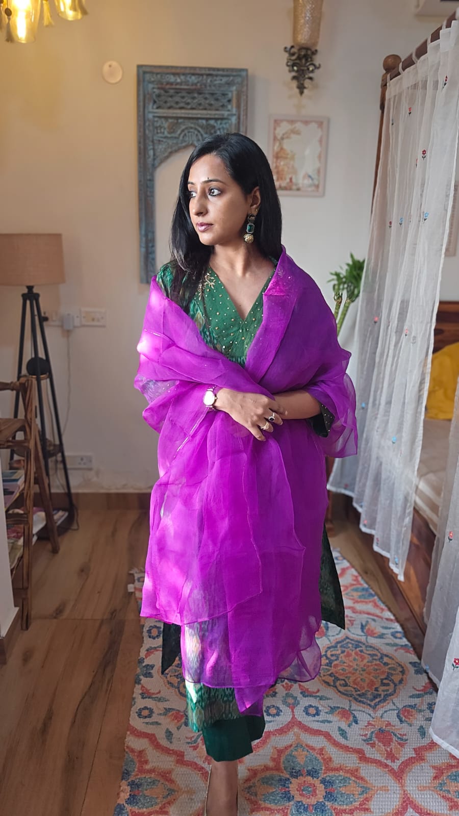 Woman wearing a green saree with a purple dupatta in a room with wooden floor and colorful rug.