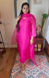 Woman in a bright pink traditional outfit standing on a patterned rug indoors.