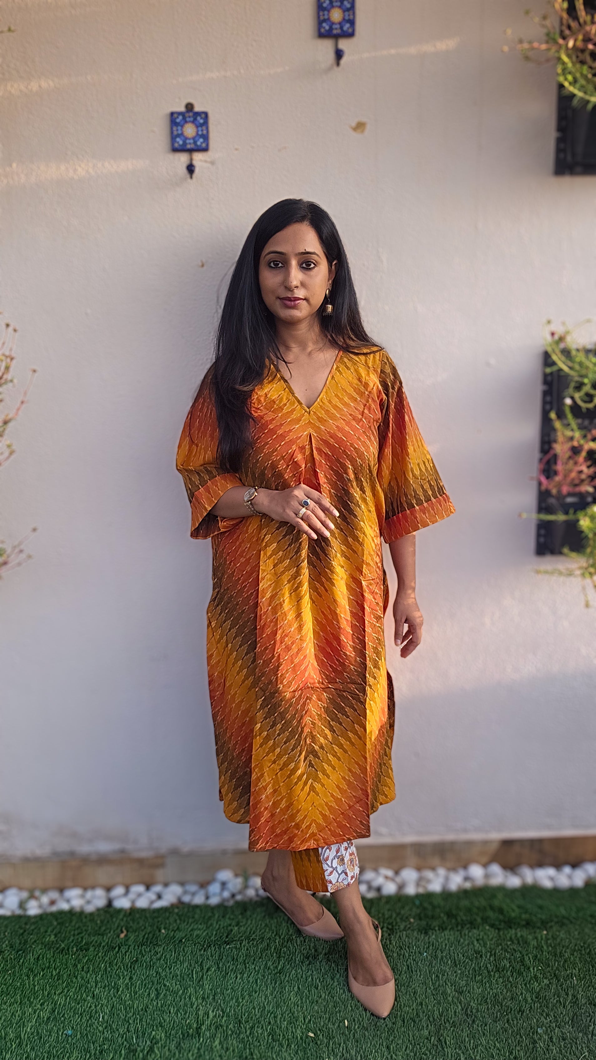 Woman wearing a colorful dress standing on grass with a white wall and plants in the background