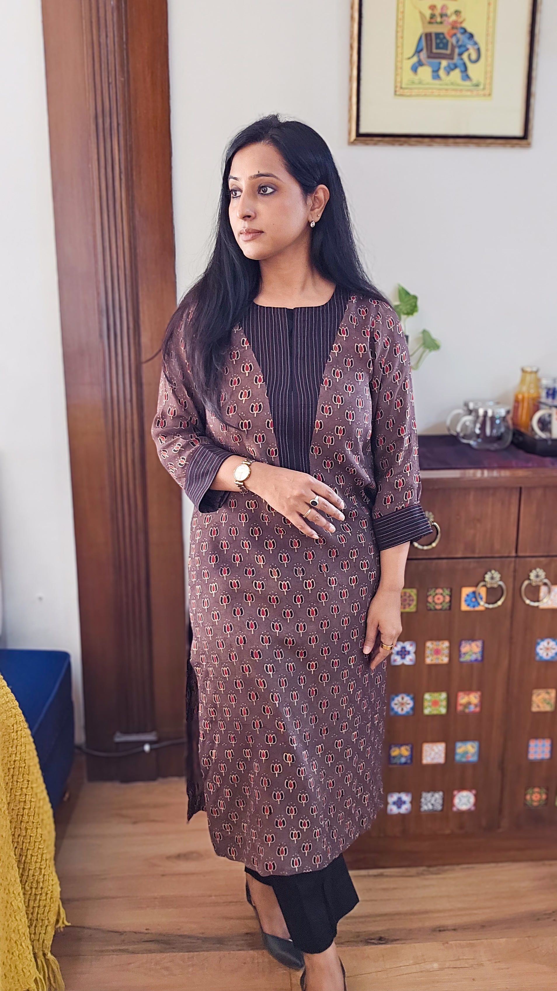 Woman wearing a patterned kurta with a matching jacket in a room with wooden furniture and decorative items.