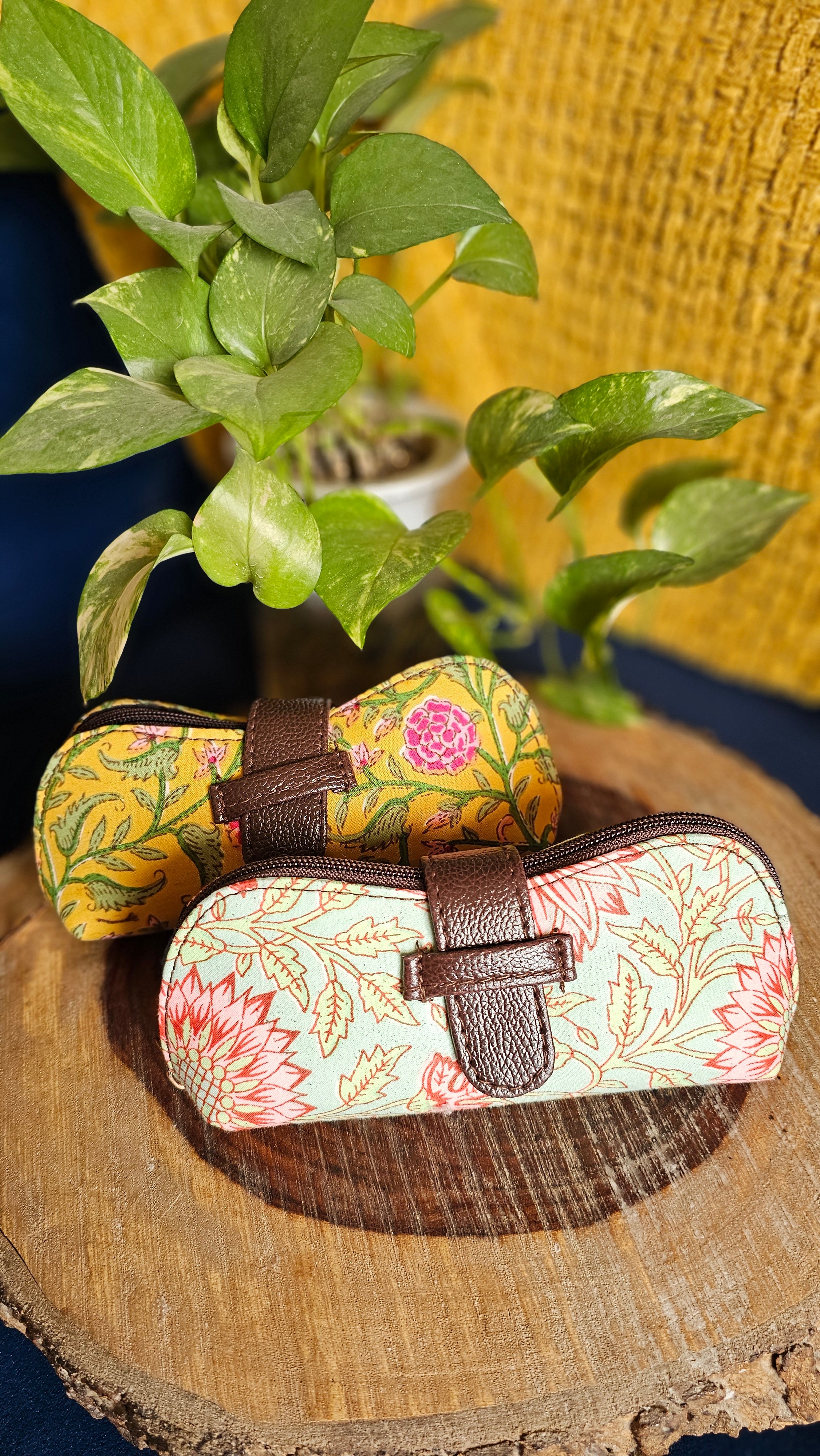 Floral-patterned sunglasses case with brown strap on a wooden surface with green leaves.