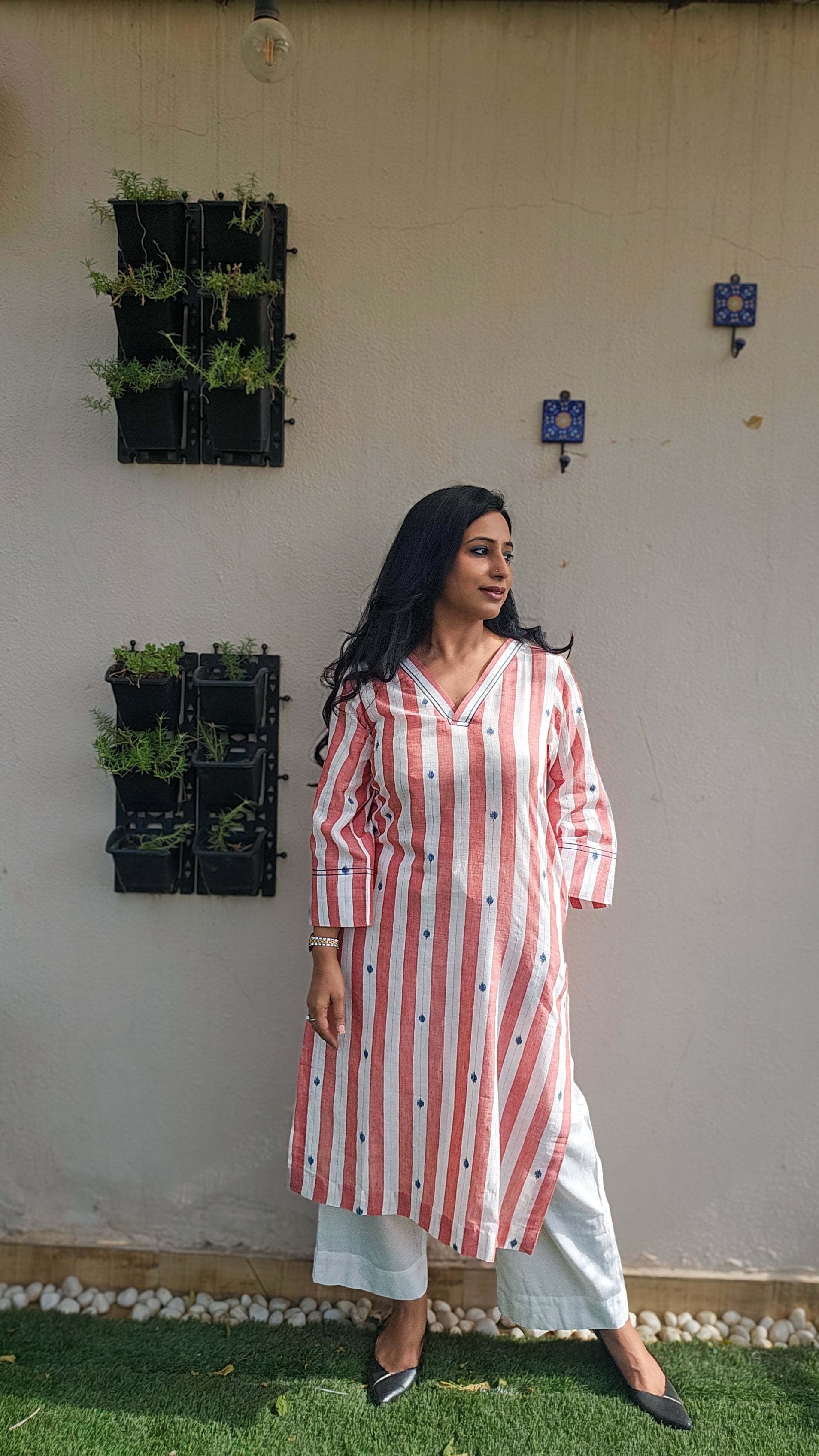 Woman wearing a red and white striped kurta standing in front of a white wall with plants.