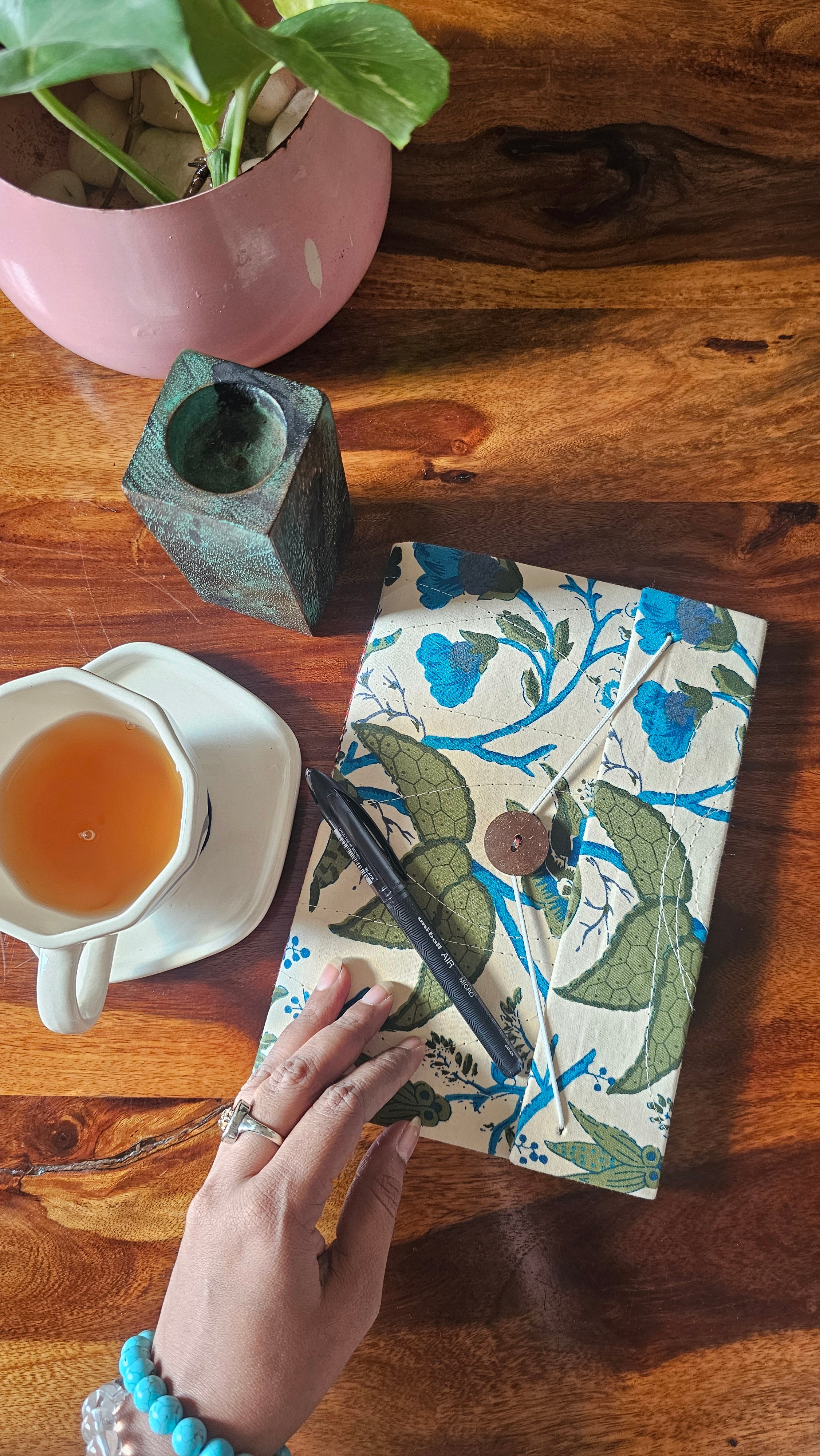 A hand holds a closed journal with a floral and leaf patterned cover, placed on a wooden table alongside a cup of tea, a small vase, and a potted plant.