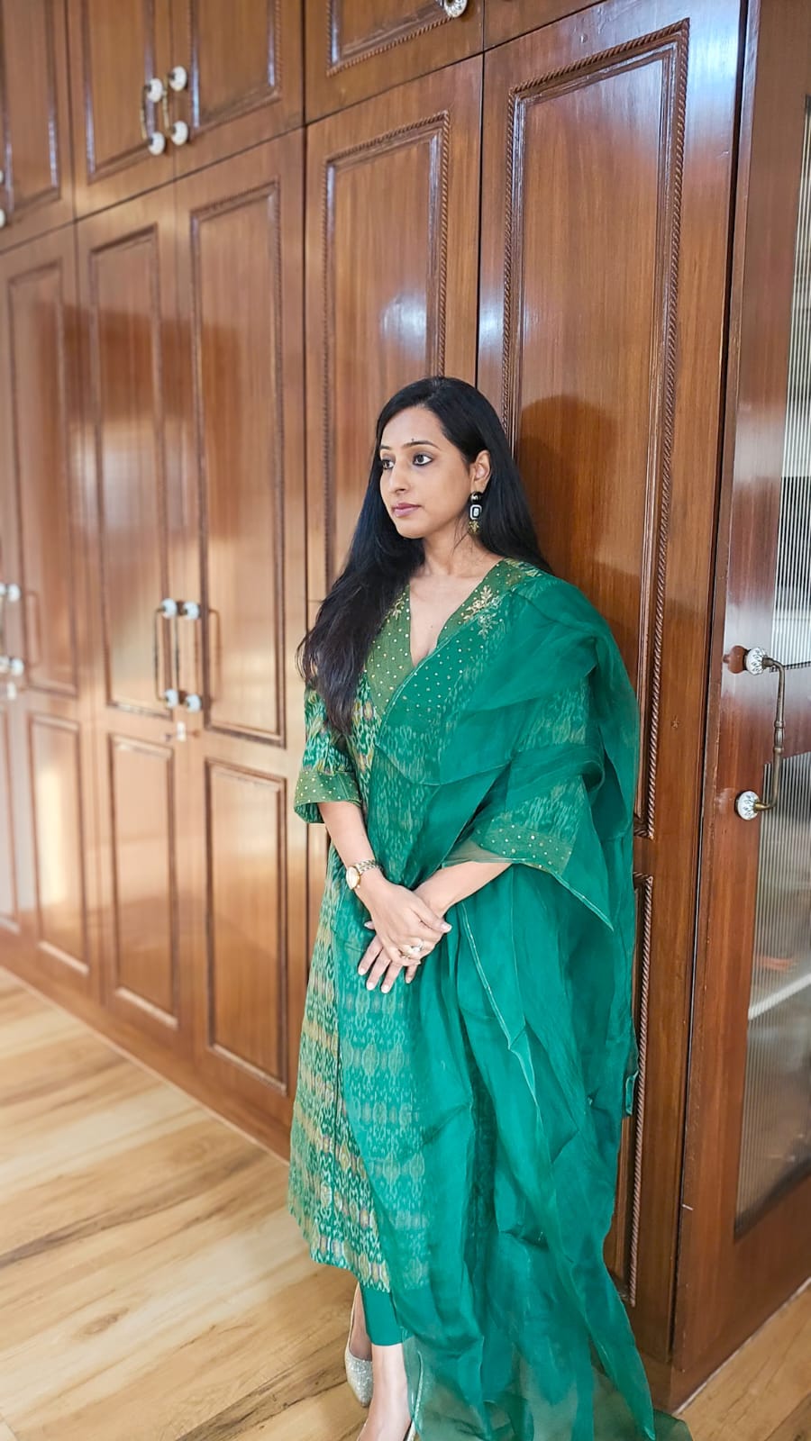 Woman in a green saree standing in front of wooden cabinets.