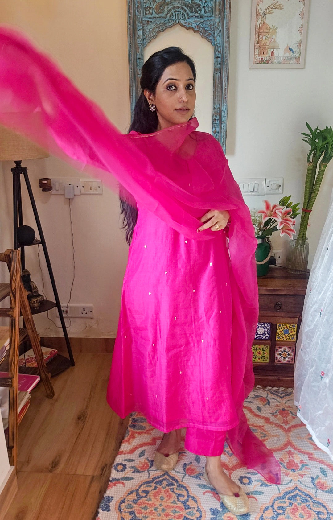 Woman in a bright pink traditional outfit  in chandedrisilk fabric with dull gold sequence work is standing in a room with wooden floor and decorative rug.