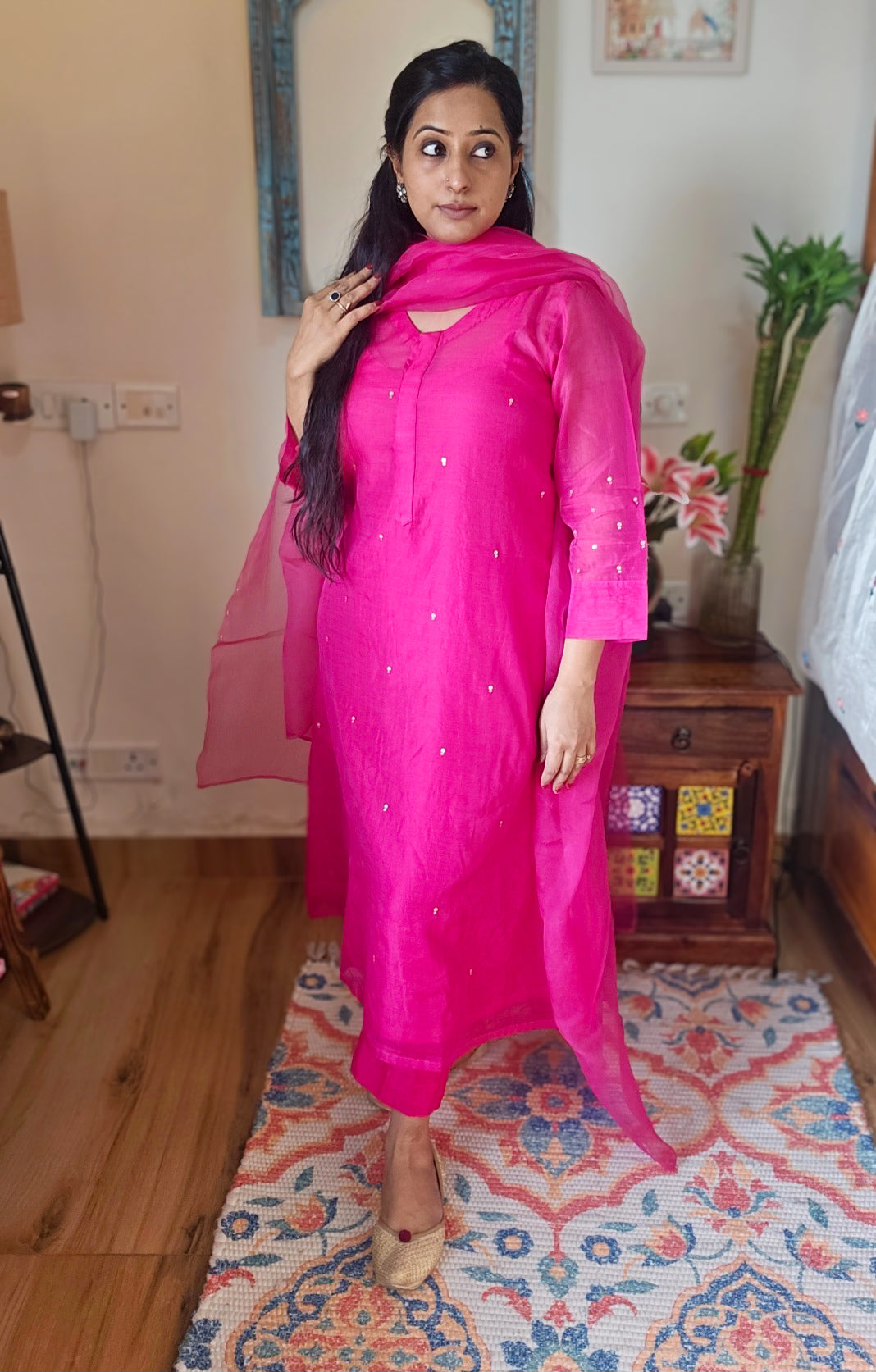 Woman in a bright pink traditional outfit standing on a patterned rug indoors.