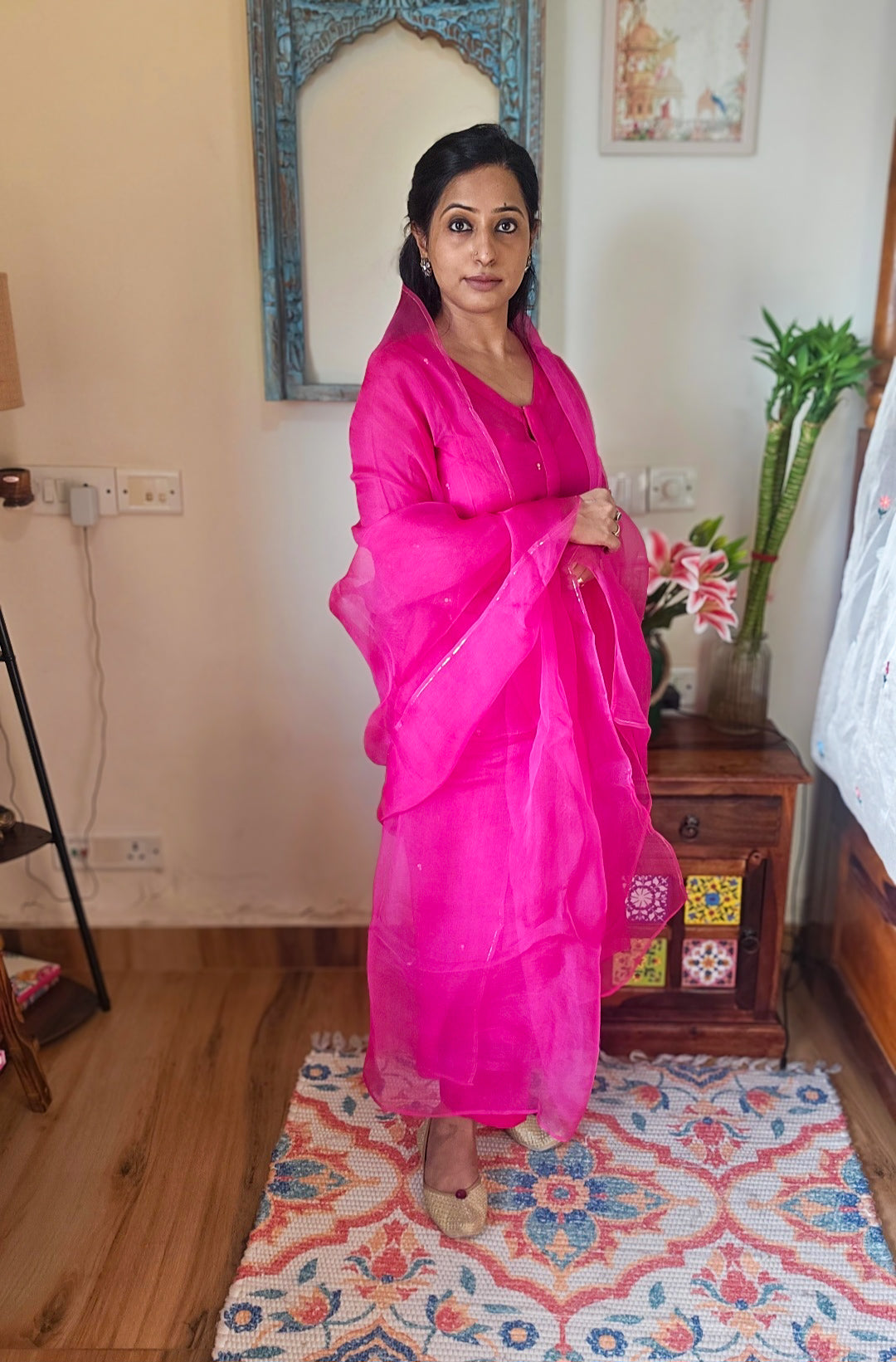 Woman in a bright pink salwar suit with organza dupatta  standing in a room with decor elements